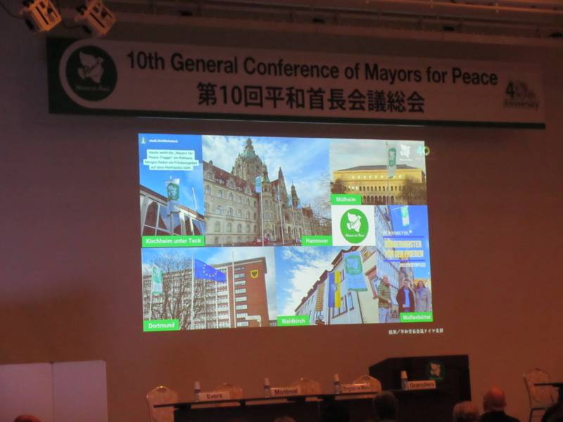 Ein Bildausschnitt von einer Präsentation, in der Fotos der Mayors for Peace-Flaggen in unterschiedlichen Mitgliedsstädten gezeigt werden.
